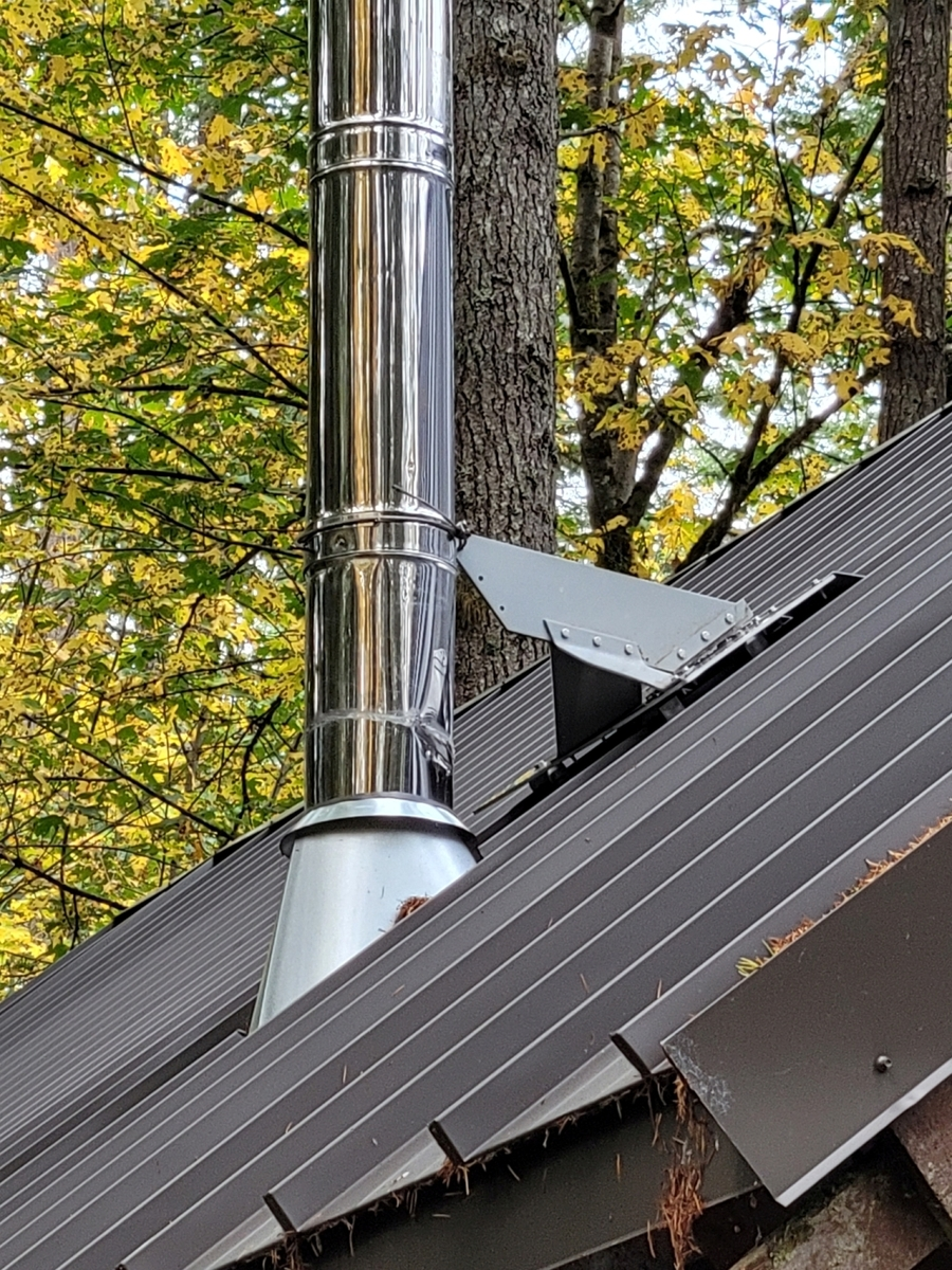 Tall metal chimney pipe extending through a standing seam metal roof with a snow-diversion protector installed upslope